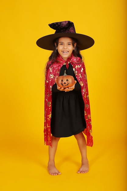 Foto una bambina con i capelli scuri con un cappello da strega e un impermeabile tiene in mano una zucca di halloween