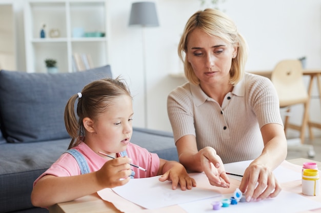 Little girl with down syndrome painting with paints together with her mother at the table at home