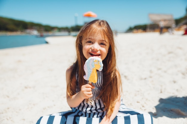 여름 드레스를 입은 다운 증후군을 가진 어린 소녀는 해변에서 아이스크림을 먹습니다