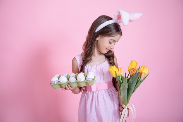 Little girl with easter bunny ears holds a bouquet of tulips and a tray of eggs in her hands on a pink studio background