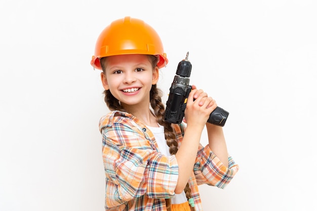 A little girl with an electric screwdriver in a protective orange construction helmet smiles broadly on a white isolated background The concept of renovation in the room of a teenage girl