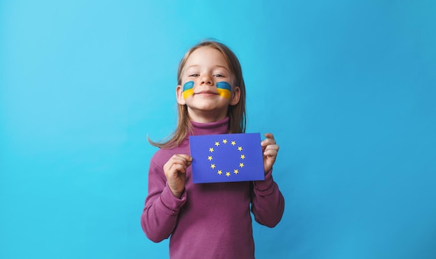 彼女の頬にウクライナの旗が描かれている少女は、孤立した青い背景に欧州連合の旗を保持しています