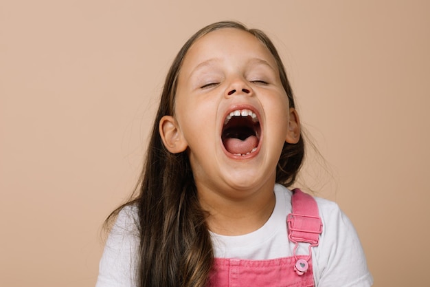 Photo little girl with fully opened mouth and closed eyes yawning looking tired wearing bright pink jumpsu...