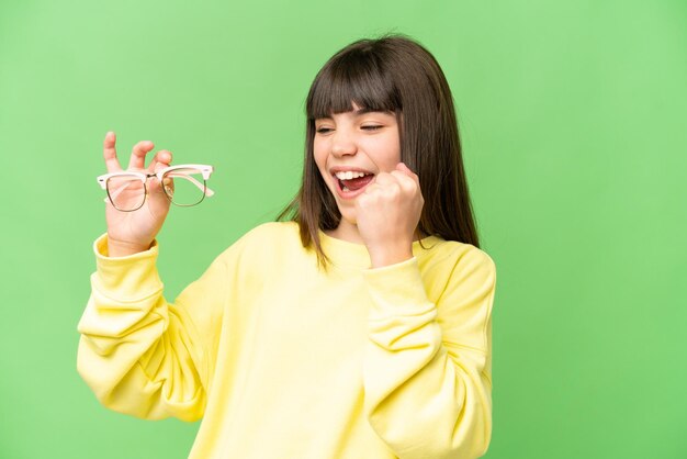 Little girl with glasses over isolated chroma key background celebrating a victory