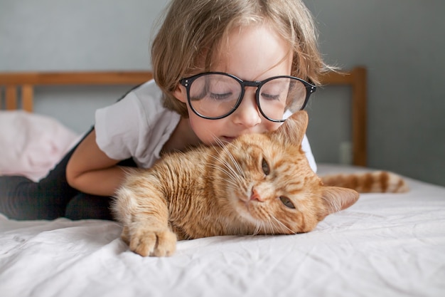 Little girl with glasses lies on the bed and hugs a fat ginger cat