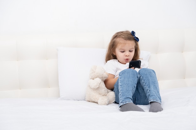 Little girl with hand in cast sitting in bed using smartphone, watching cartoon or education video.