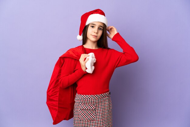 Foto bambina con cappello e sacco di natale isolato su sfondo viola avendo dubbi