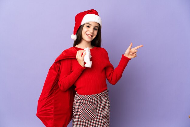 Bambina con cappello e sacco di natale isolato sulla parete viola che punta il dito a lato