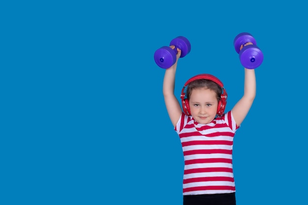 A little girl with headphones is playing sports with dumbbells in her hands on blue background