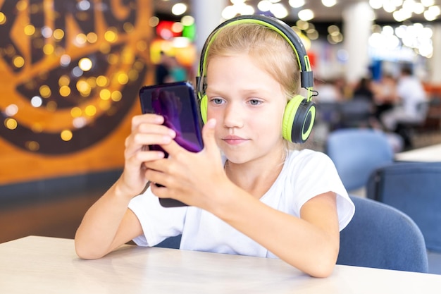 A little girl with headphones is watching cartoons on her phone sitting at a table in a cafe Cute schoolgirl listening to music