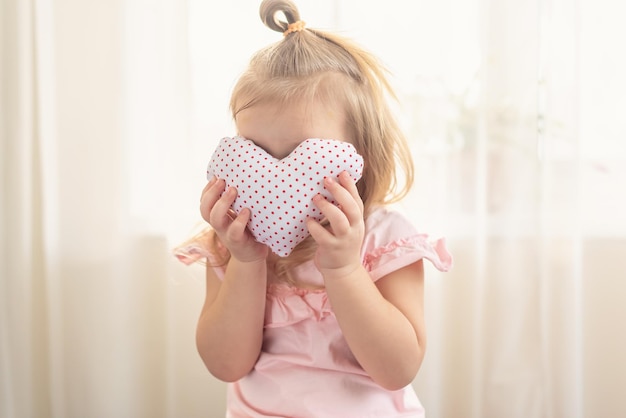 Little girl with heart shaped pillow indoor, hiding behind heart. Concept of love, happiness, kindness, gift, handmade Valentine, familly. Celebrating love day, women's day, World Heart Day, birthday.