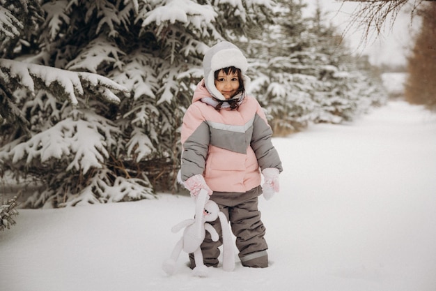 A little girl with a knitted hare stands on the street under the flying snow. winter day