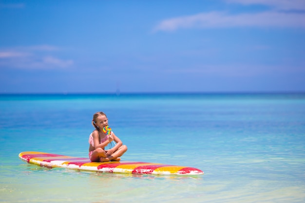 Little girl with lollipop have fun on surfboard in the sea