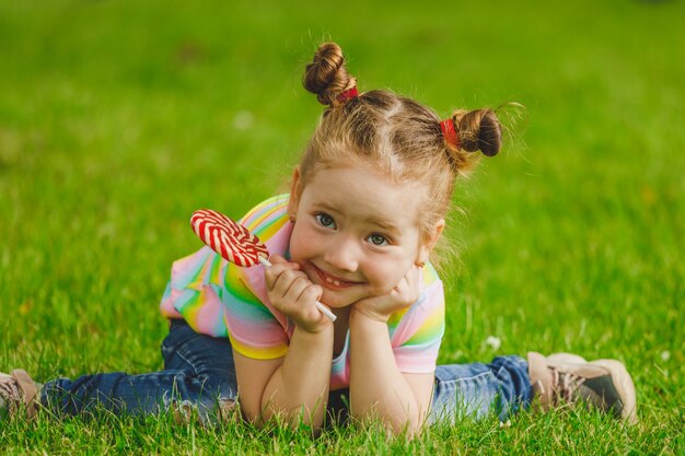 Little girl with lollipop sits on the grass in summer in the park