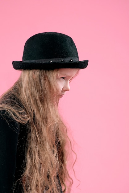 little girl with long blonde hair in black blazer and stylish black hat on pink background