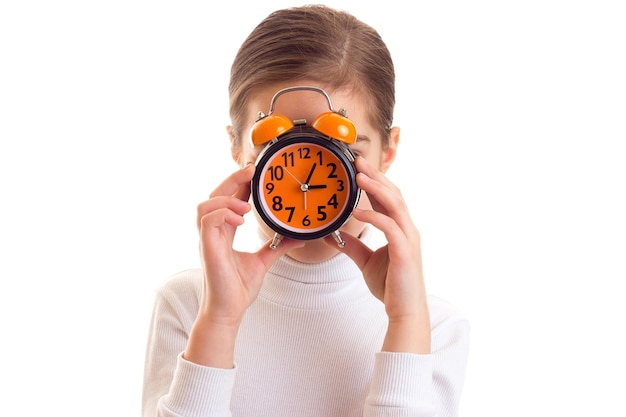 Little girl with long chestnut ponytail hiding her face beyond orange clock on white background