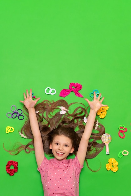 A little girl with long hair lies with elastic bands and hair clips and accessories Hairstyles for girls Green isolated background Copy space Space for text
