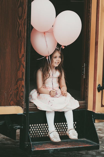 A little girl with long hair in a pink dress gets out of the tram with balloons in her hands. High quality photo