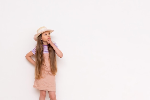 A little girl with long hair in a summer sundress and a straw hat leans against a white walls and looks at your advertisement smilingCopy space