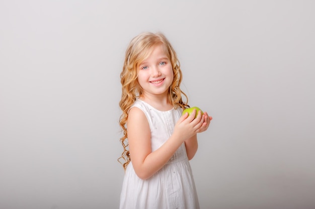 A little girl with long hair and a white dress holds an apple on a white background