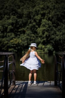 Una bambina con i capelli lunghi in abito bianco si trova sulla riva del fiume. passeggiata in riva al lago in una giornata di sole estivo