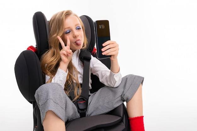 A little girl with makeup and long blonde hair sitting in a car baby chair with mobile phone, do selfie and shows peace