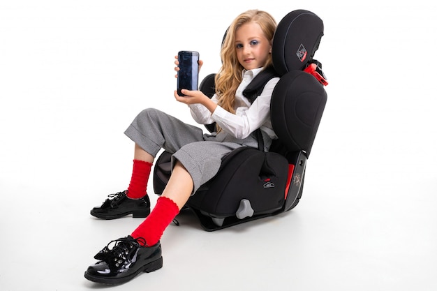 A little girl with makeup and long blonde hair in a white shirt, red pull-ups, pants in a cage, red socks and shoes with phone in a baby chair