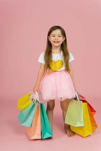 Little girl with many shopping bags on pastel pink wall. Pretty joyful young girl in tulle skirt, with long blonde hair walking with colorful packages on pink wall