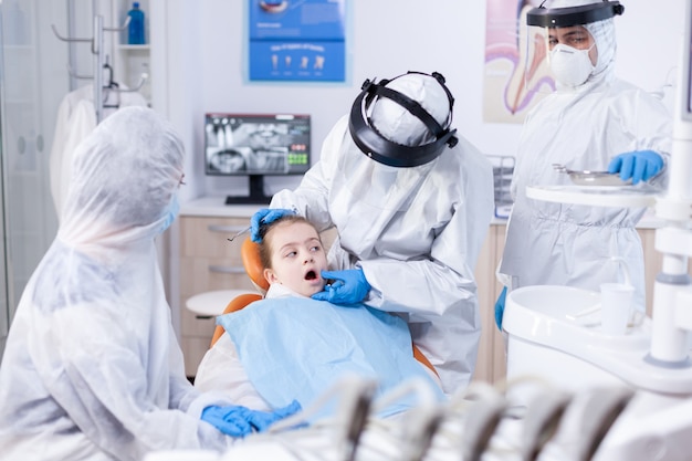 Little girl with mouth open sitting on dental chair dressed in ppe uit as safety precaution in the course of coronavirus. Dentist in coronavirus suit using curved mirror during teeth examination of ch