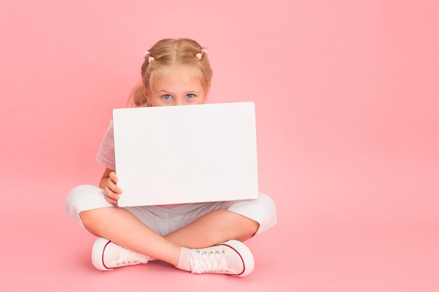 Little girl with notebook sitting with legs crossed on pink background with copy space