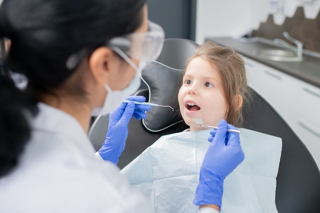 Little girl with open mouth looking at the dentist in protective workwear and gloves going to examine oral cavity