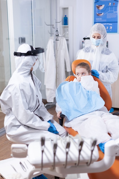 Little girl with painfull expression sitting on dental chair wearing bib. Stomatologist during covid19 wearing ppe suit doing teeth procedure of child sitting on chair.