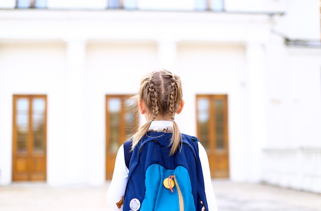 Little girl with pigtails braids in uniform dress goes to school with blue backpack