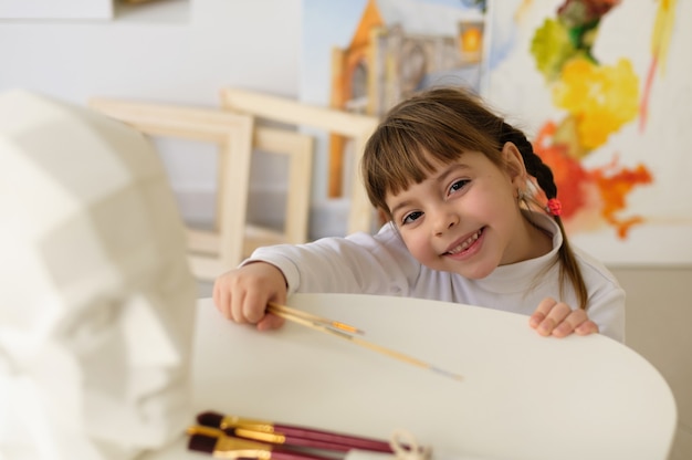 Little girl with pigtails in a home studio-workshop holds art brushes and smiles