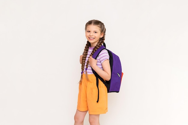 Маленькая девочка с косичками и шортами со школьным рюкзаком на спине на белом изолированном фоне Подготовительные летние курсы для детей