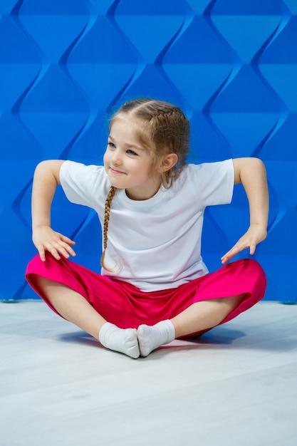 Little girl with pigtails in a white T-shirt on a blue background. She sits in a lotus position on the floor and smiles and waves her hands.