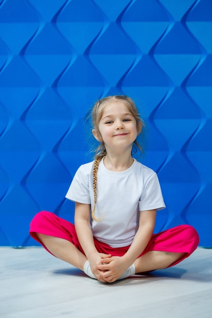 Little girl with pigtails in a white t-shirt on a blue background. she sits in a lotus position on the floor and smiles and waves her hands