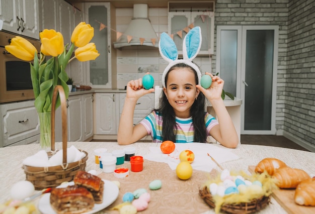 A little girl with pink bunny ears holds easter eggs on her face near her eyes in the kitchen