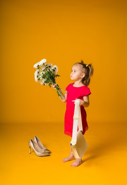 Little girl with ponytails in a red dress stands sideways with a bouquet of white flowers on a yellow surface with space for text