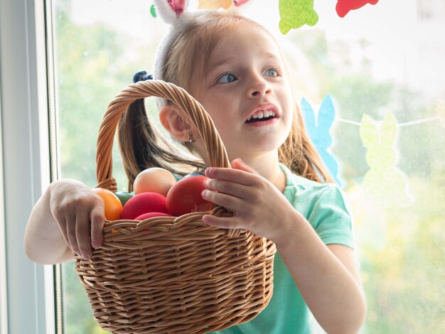 A little girl with rabbit ears is sitting on the windowsill, holding a basket of Easter eggs