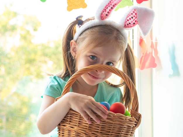 A little girl with rabbit ears on the windowsill smiles with a basket of Easter eggs