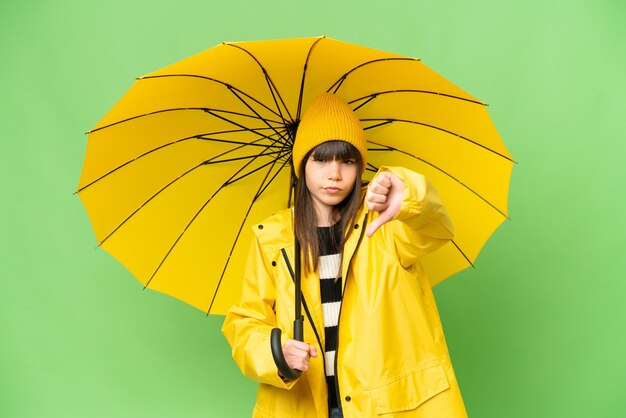 Little girl with rainproof coat and umbrella over isolated chroma key background showing thumb down with negative expression