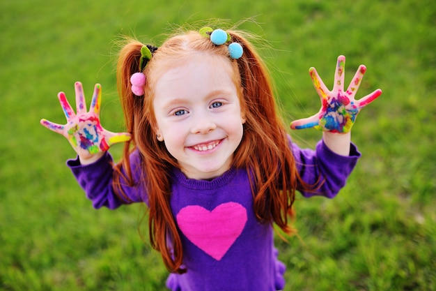 Little girl with red hair shows her hands dirty with multicolored paints and smiles