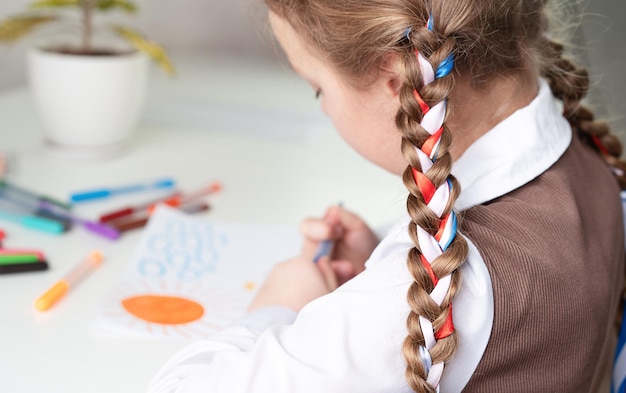 A little girl with ribbons in her hair is sitting at a table and drawing