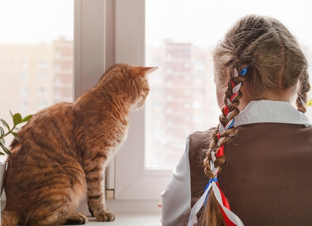 Little girl with ribbons in her hair look out the window with a cat