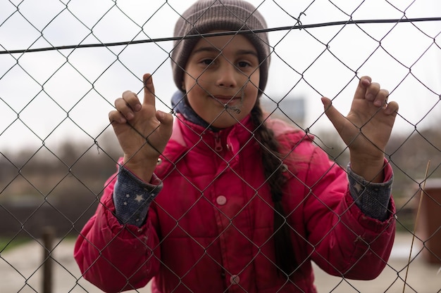 Foto una bambina con uno sguardo triste dietro una recinzione metallica. problema sociale dei rifugiati e dei migranti forzati.