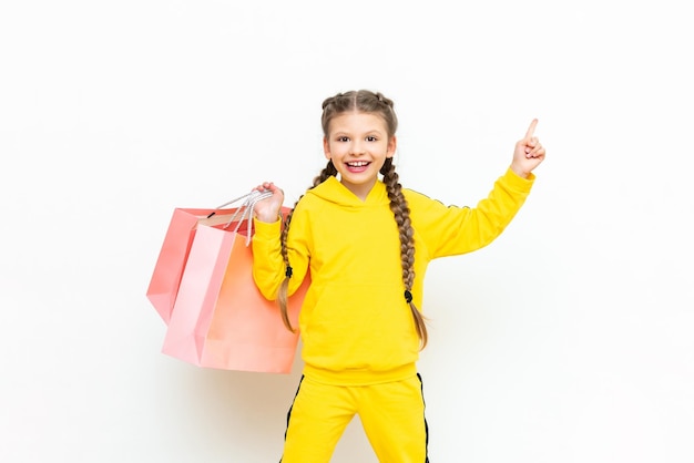 Una bambina con lo shopping in una tuta sportiva gialla guarda il tuo annuncio su uno sfondo bianco isolato un bel bambino è felice con i nuovi acquisti