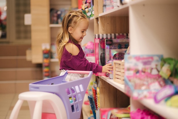 Little girl with small shopping cart in kids mall happy girl choosing what to buy in toy store