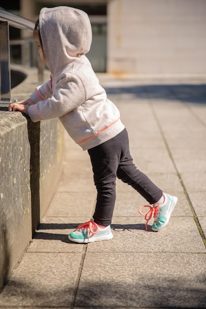 Bambina con scarpe da ginnastica e felpa con cappuccio che si scalda prima dell'allenamento in una piazza della città