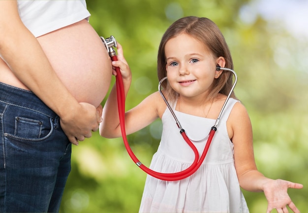 彼女の妊娠中の母親の腹を調べる聴診器を持つ少女